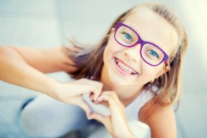Smiling little girl with dental braces and glasses showing heart with hands